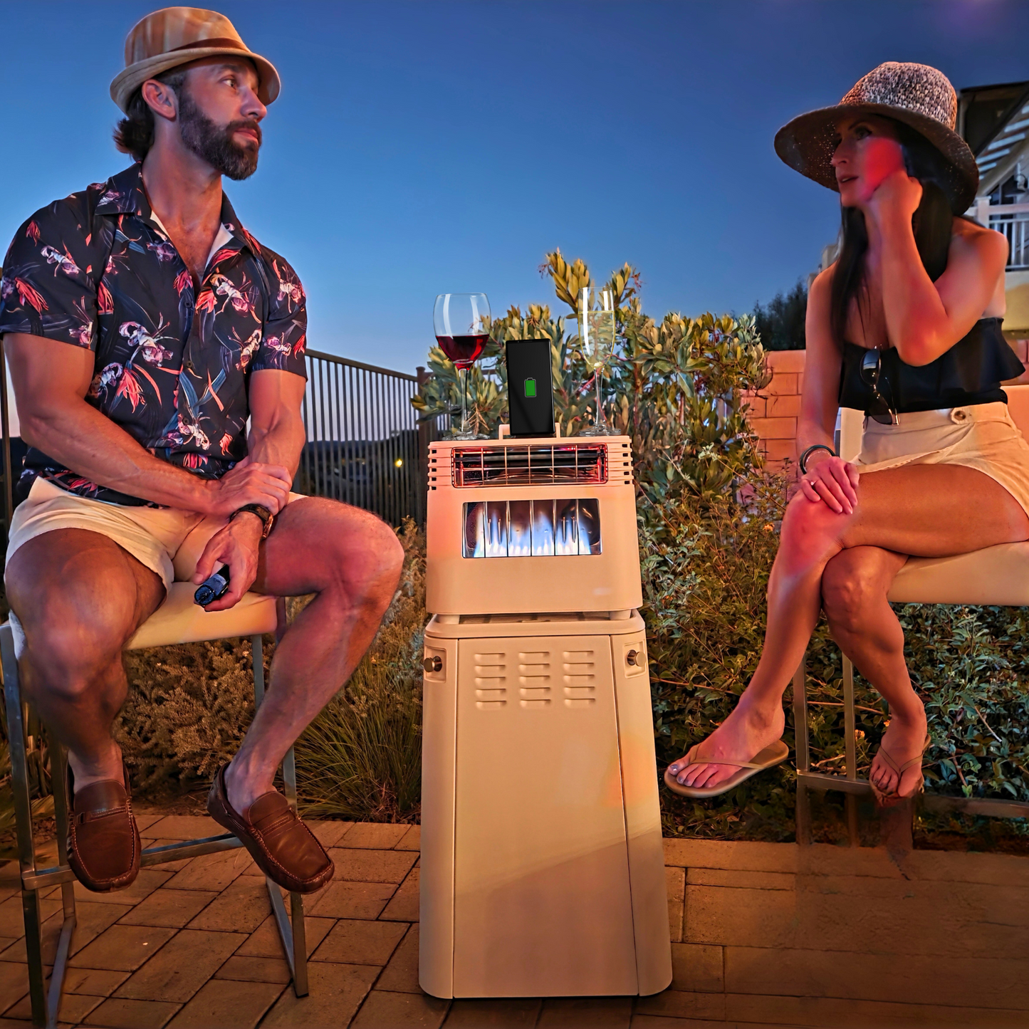 Two people sitting on chairs with a portable air conditioner between them outdoors at night.