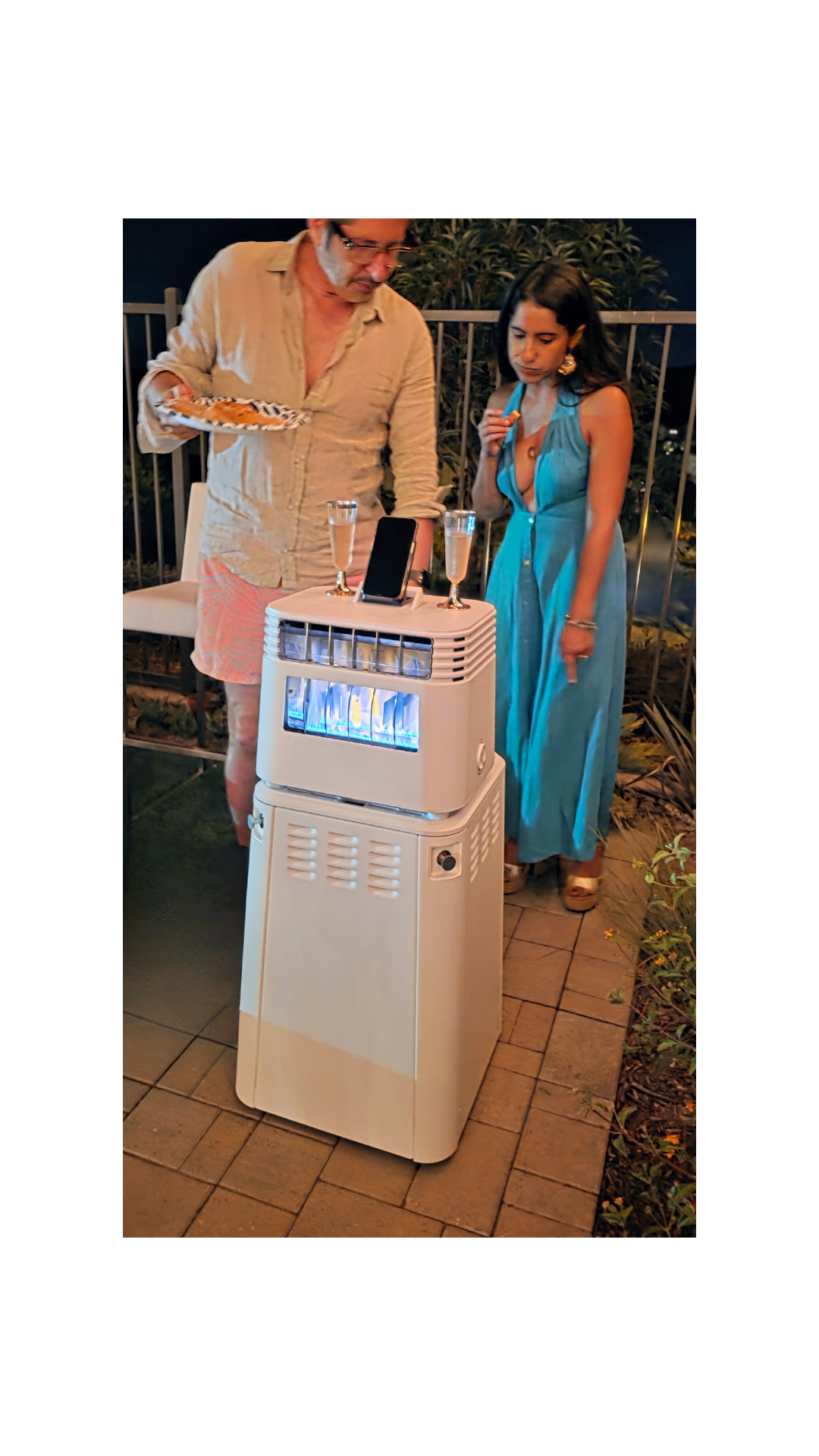Man and woman standing near white outdoor heater with smartphone and two glasses on top at night