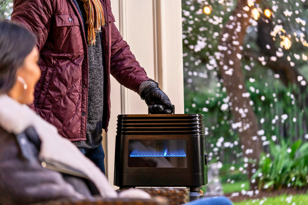 Person in winter jacket and gloves holding portable outdoor propane heater with visible blue flame near seated woman in cold garden setting