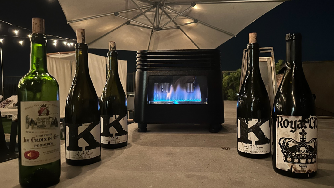 Six wine bottles arranged around a small black tabletop propane heater under a patio umbrella at night