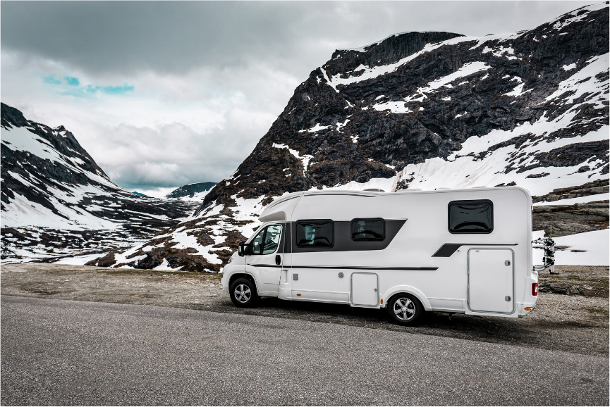 White camper van parked on mountain road with snow-covered rocky peaks under cloudy sky