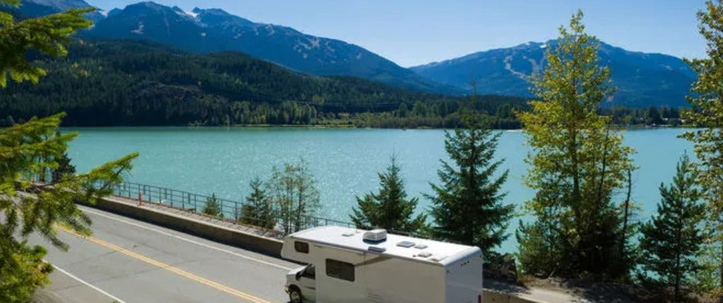 White RV parked by road alongside large blue lake surrounded by pine trees and mountains under clear sky
