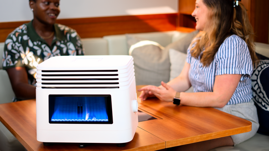 Modern white heater with blue flame on wooden table, people chatting in cozy room