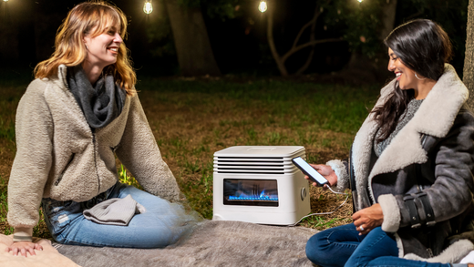 Two women sitting outdoors on a blanket at night enjoying warmth from a portable electric heater