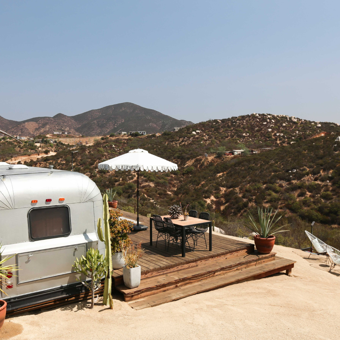 Outdoor wooden deck with table, chairs, white umbrella, and silver caravan in desert landscape