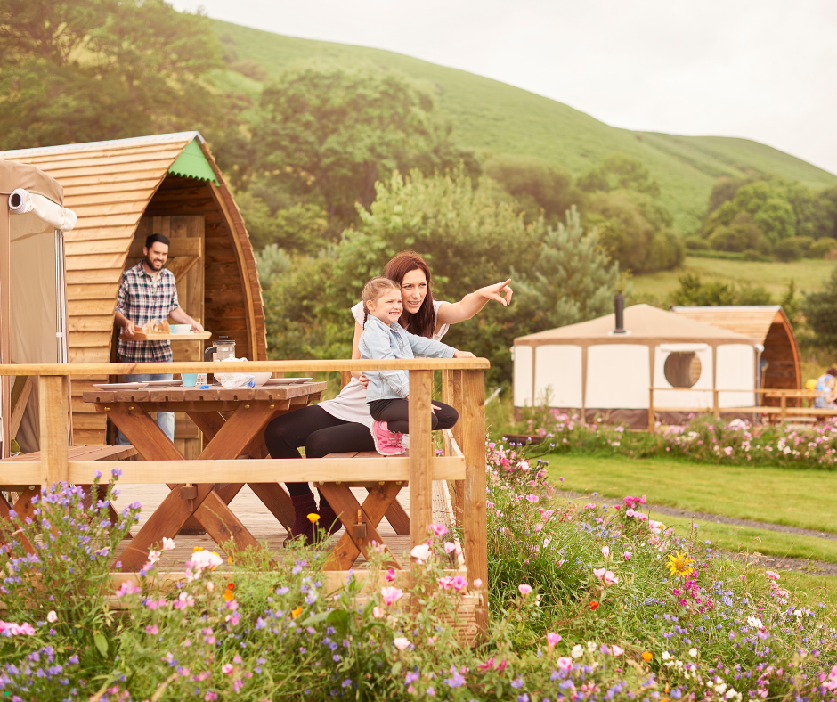Mother and daughter sitting on wooden deck of glamping pod, man carrying breakfast tray, wildflowers and green hills