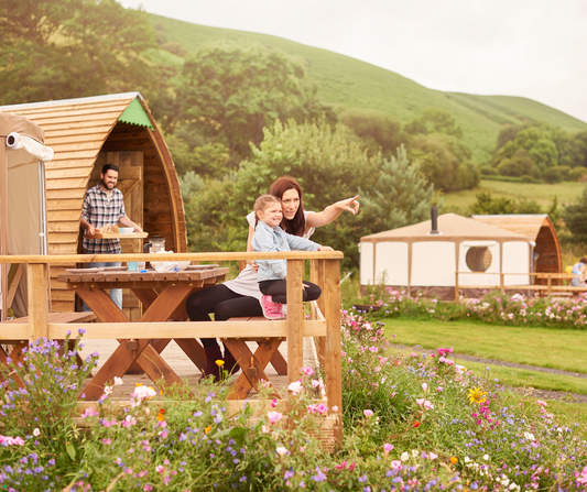 Mother and daughter sitting on wooden deck of glamping pod, man carrying breakfast tray, wildflowers and green hills