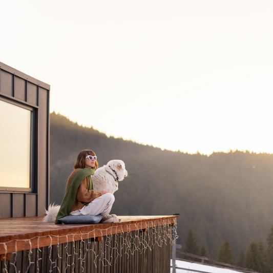 Woman in sunglasses sitting on wooden deck with white dog overlooking a forested mountain at sunset