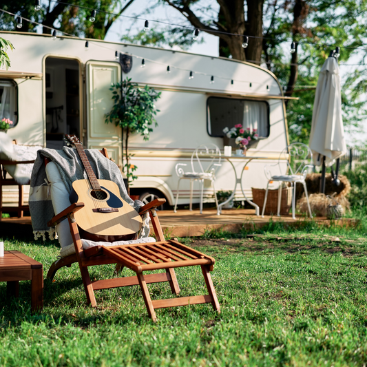 Wooden lounge chair with guitar and blanket on grass beside white camper and outdoor patio setup