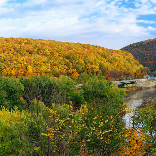 Scenic autumn landscape with colorful trees on hills and a river with a bridge under a blue sky
