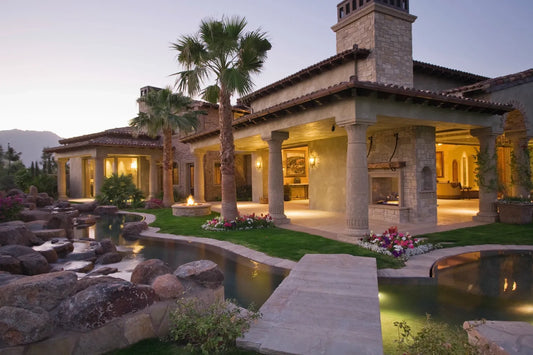 Luxury villa with stone pillars, outdoor fireplaces, palm trees, garden, and a winding water feature at dusk