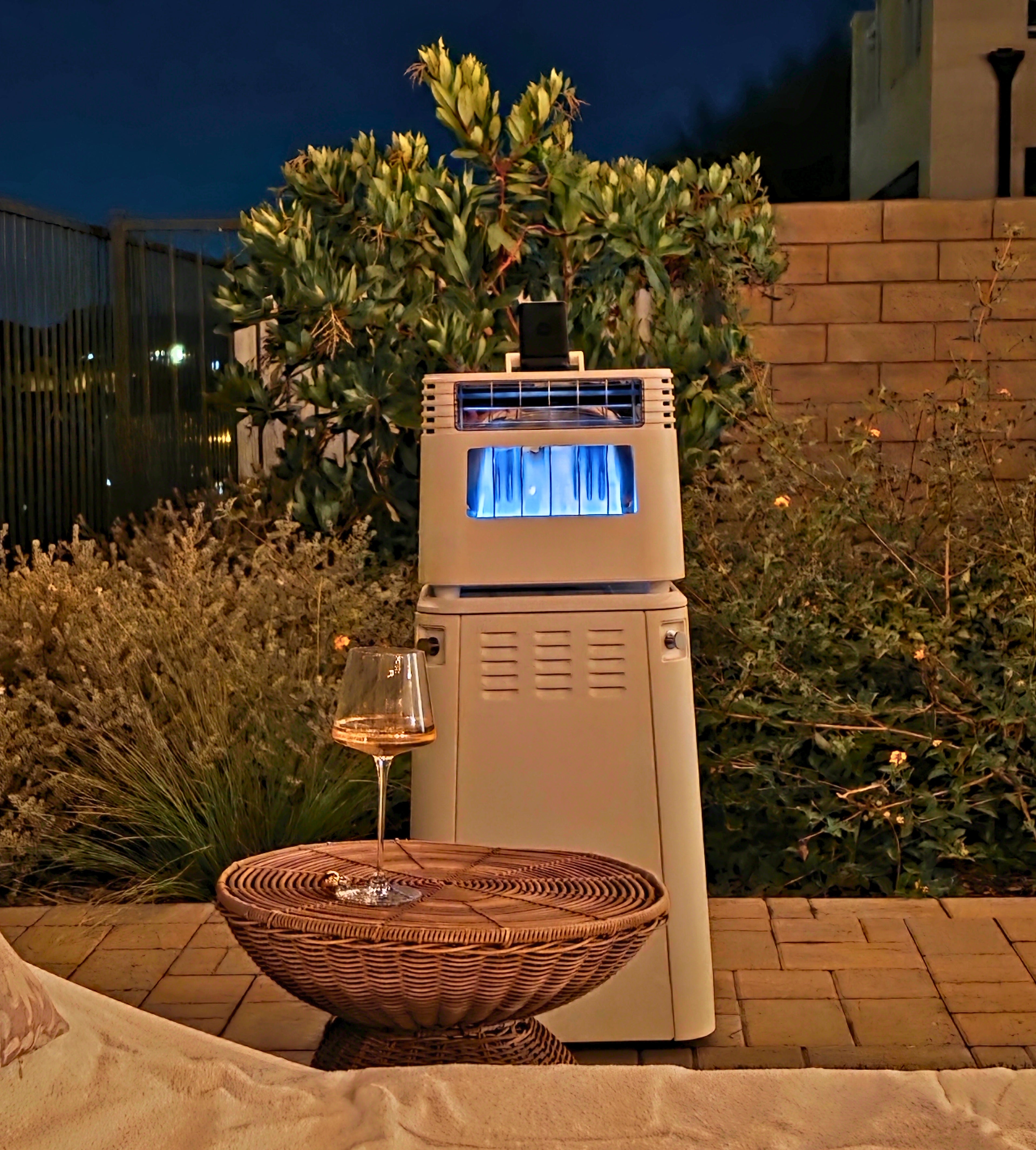 Outdoor patio gas heater with blue flame near wicker table holding a glass of wine at night